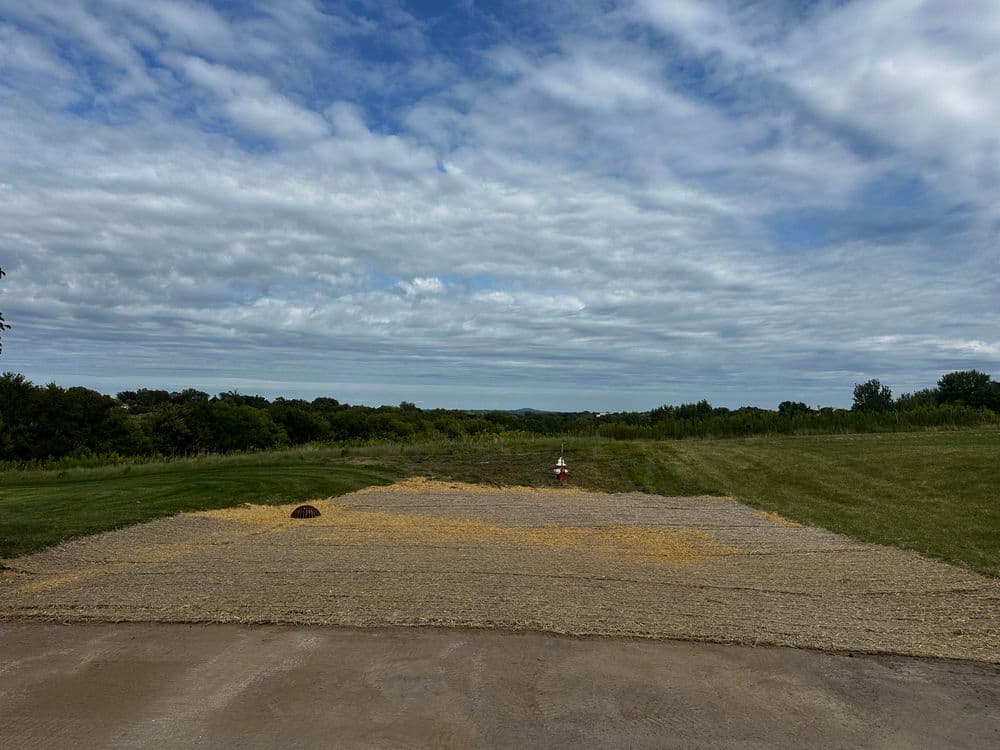 Cleared land with straw cover under a cloudy sky, surrounded by greenery and trees.