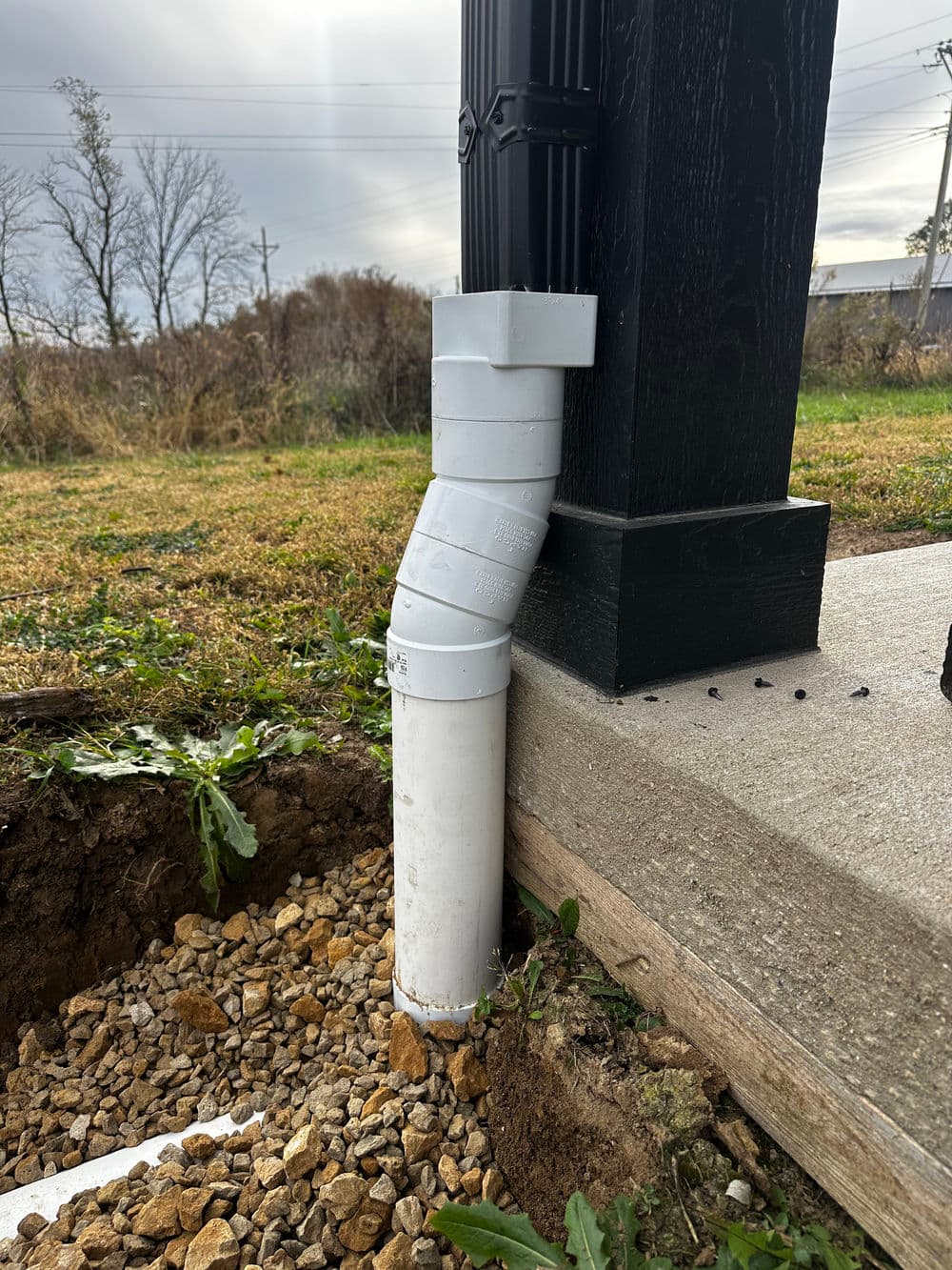 White drainage pipe connected to a black post, positioned on a gravel base outdoors.
