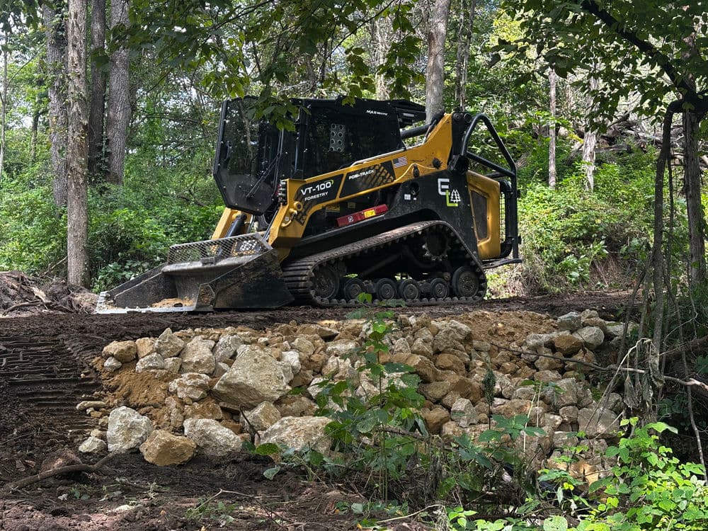 Yellow compact track loader working on a hillside with rocks and trees in the background.