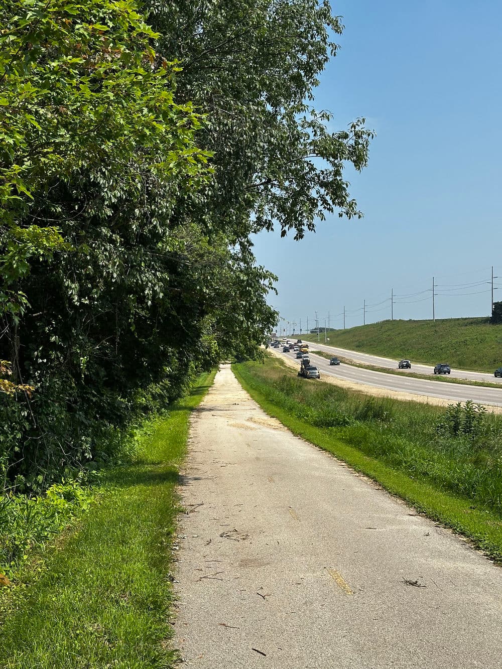 Paved walking path beside a busy highway under clear blue sky and green trees.