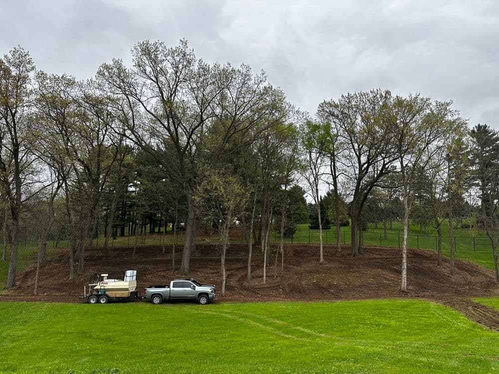 Truck and trailer on cleared land surrounded by trees and grassy area under overcast sky.
