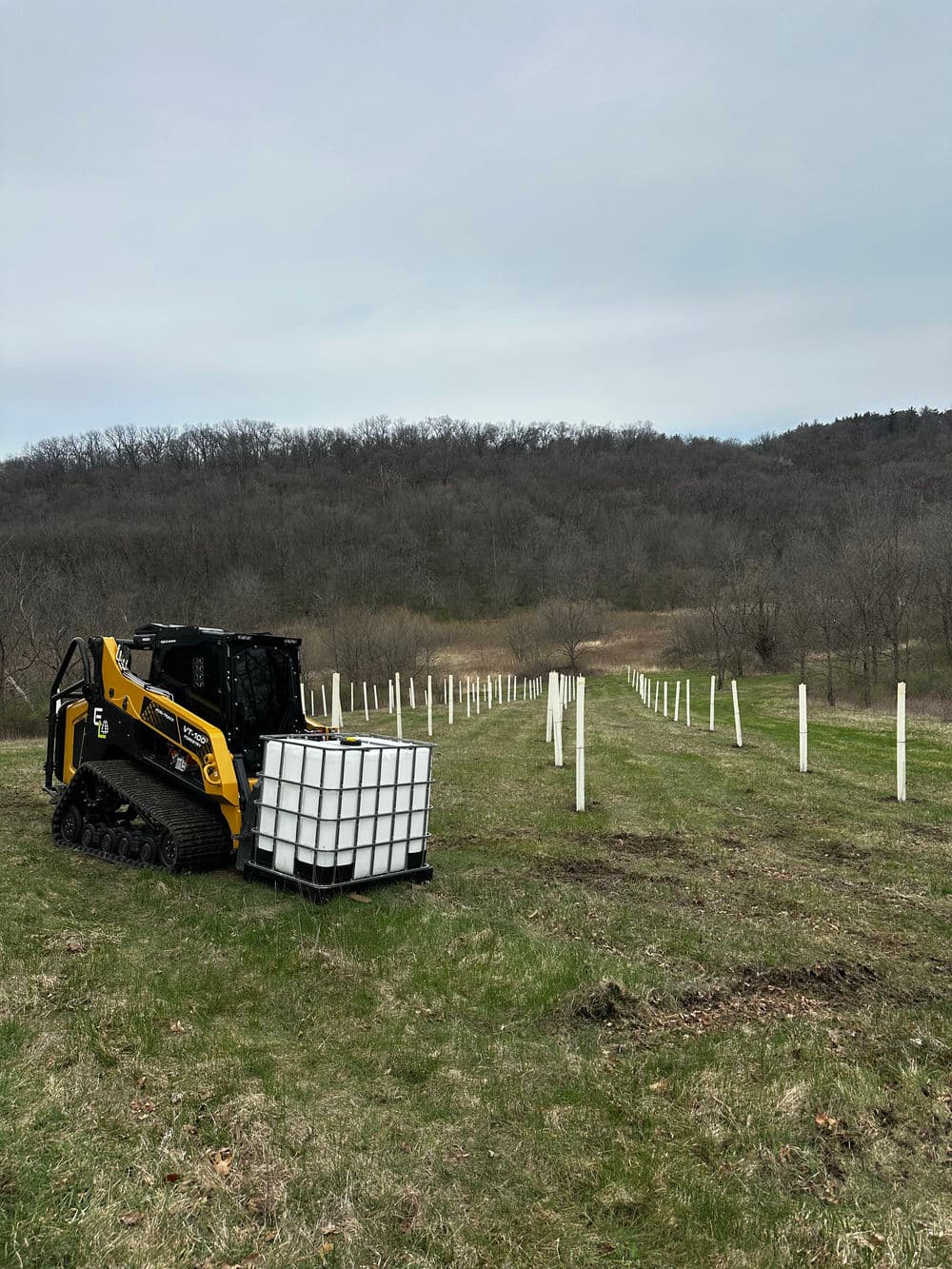 Skid steer with a large water tank in a vineyard with posts and hills in the background.