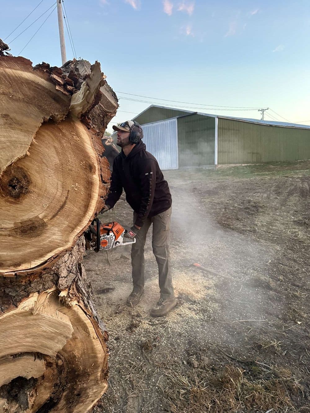Man using a chainsaw on a log with smoke and a barn in the background during daylight.